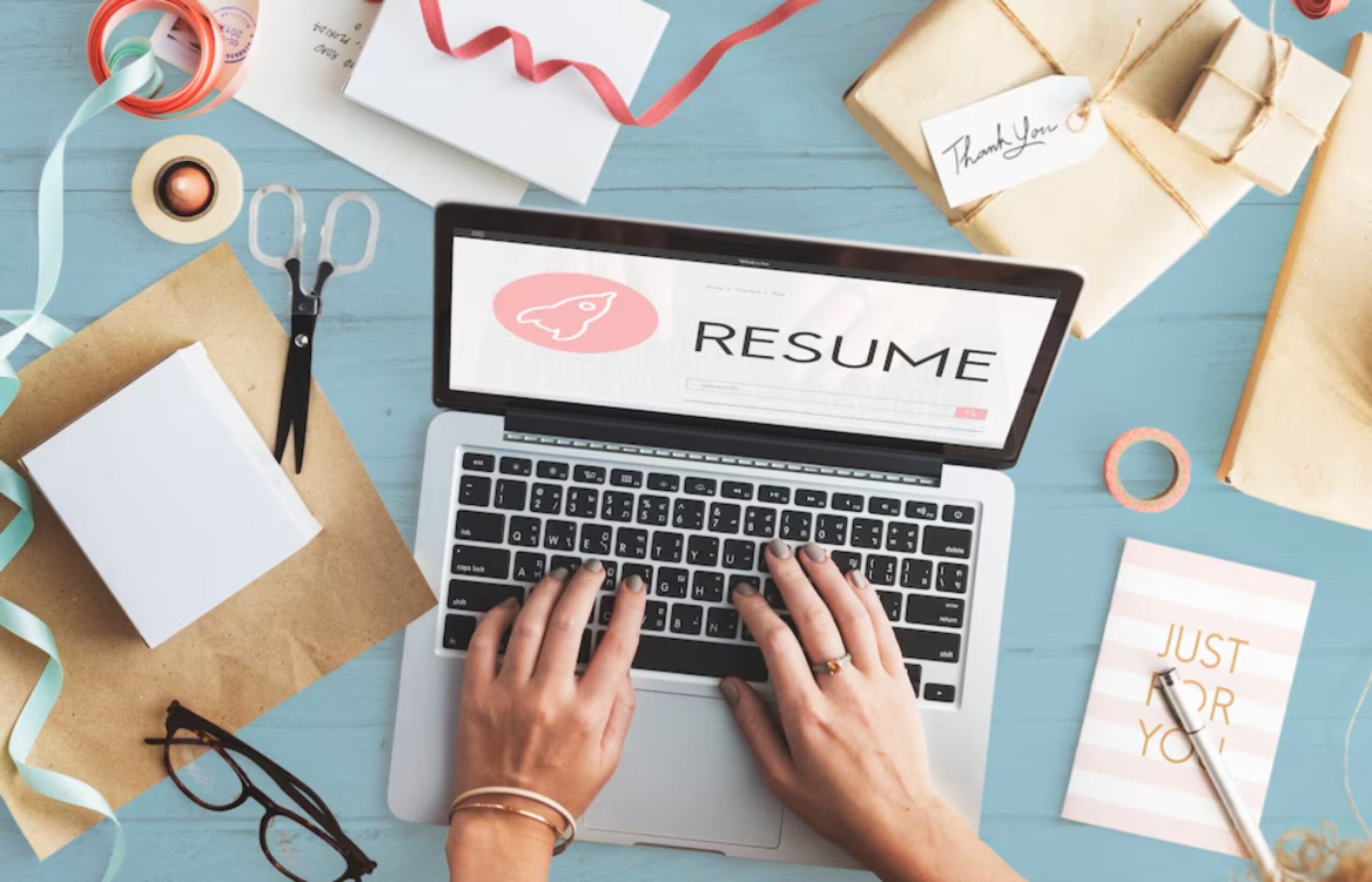 A person types on a laptop displaying a resume, surrounded by scissors, gift boxes, ribbons, and stationery on a blue table.