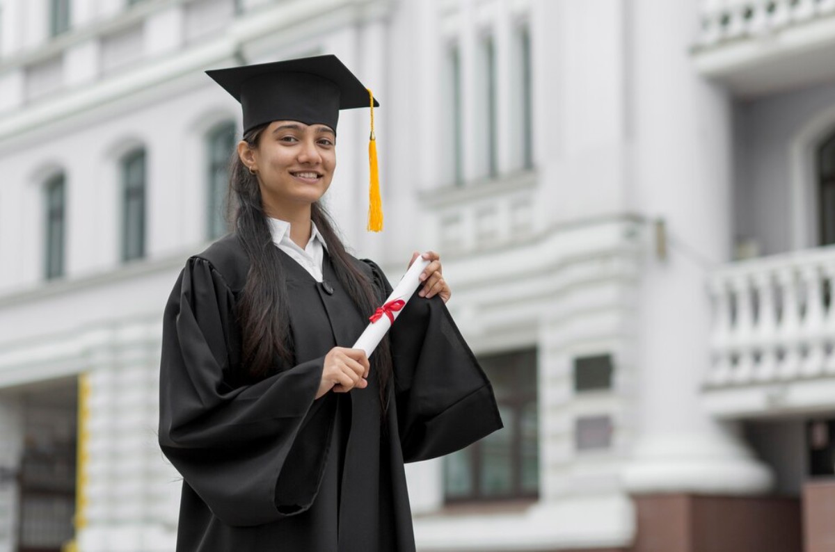 A graduate in a black gown and cap holds a diploma with a red ribbon, posing confidently in front of an elegant building.