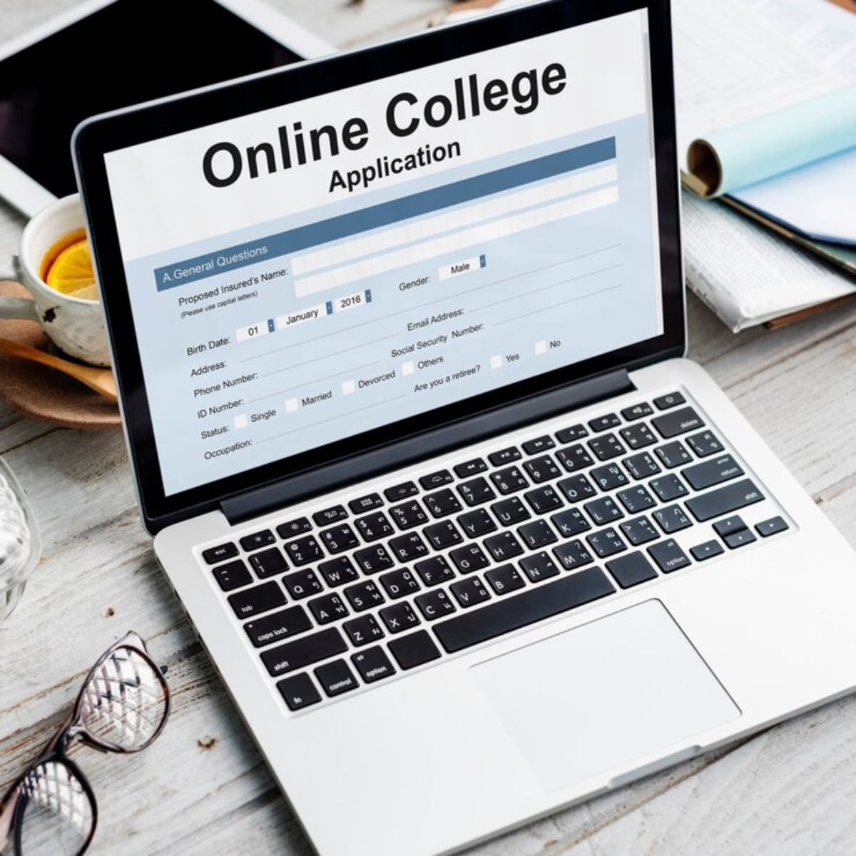 A laptop displaying an online college application form, surrounded by glasses, tea, and notebooks on a wooden table.