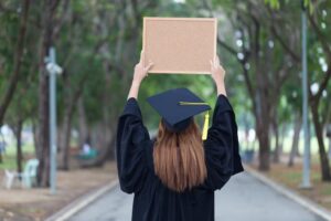A woman wearing a graduation gown and cap stands outdoors, holding up a board. In the background, there is a tree.