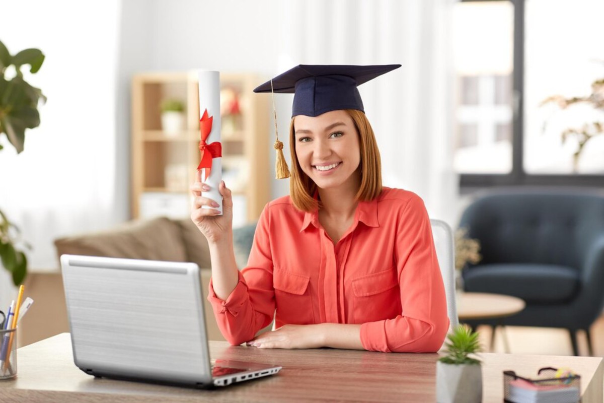 A graduate in a cap and gown holds a diploma with a red ribbon, seated at a desk with a laptop and a plant.